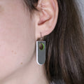 Close-up of an ear wearing a unique arch shaped linoleum earring, grey with white outline and a green cutout circle. on a small hoop