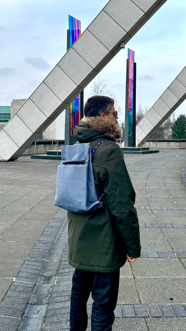 Person wearing a green coat and carrying a blue backpack in an urban setting with modern architecture.